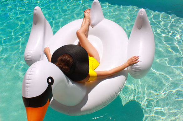 Kelly Golightly, a woman wearing a yellow one piece bathing suit and a black sunn hat, floats on a giant white swan pool float in an aqua pool.