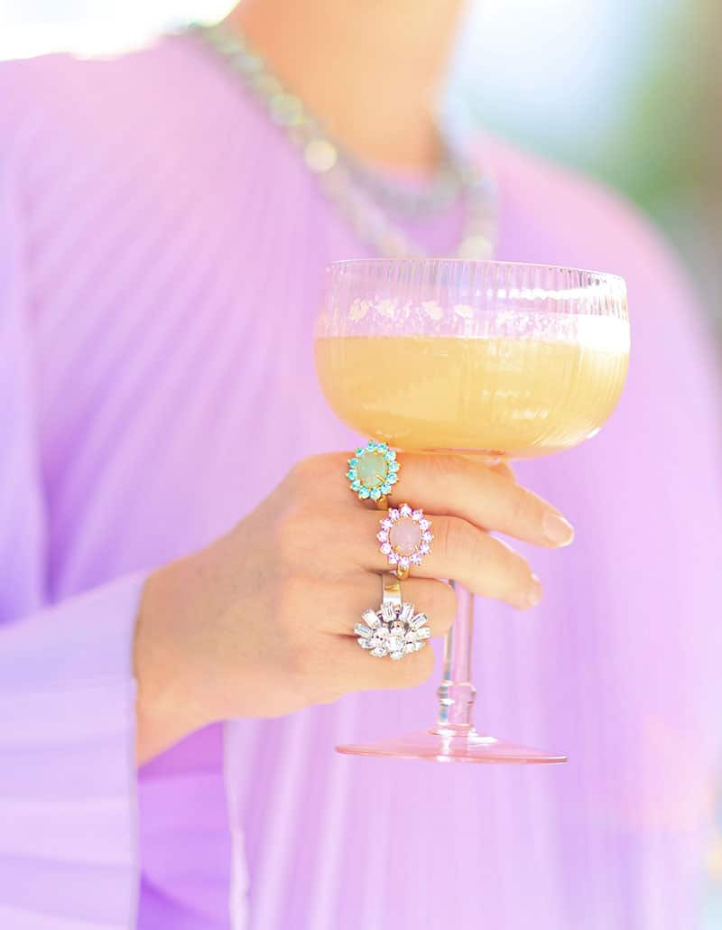 Hand holding a pink coupe cocktail glass wearing three candy-colored cocktail rings in Palm Springs style
