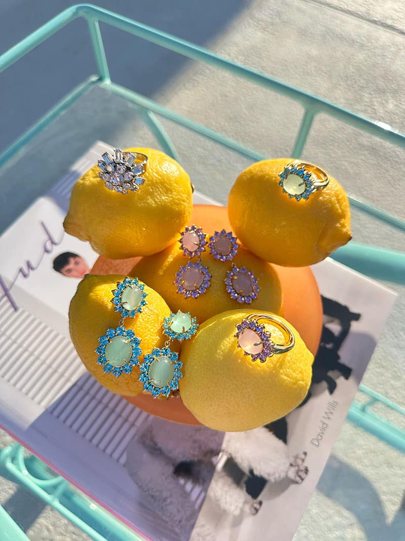 Flat lay of Palm Springs-inspired poolside jewelry including statement candy-colored earrings and cocktail rings resting on lemons atop a vintage aqua bar cart 